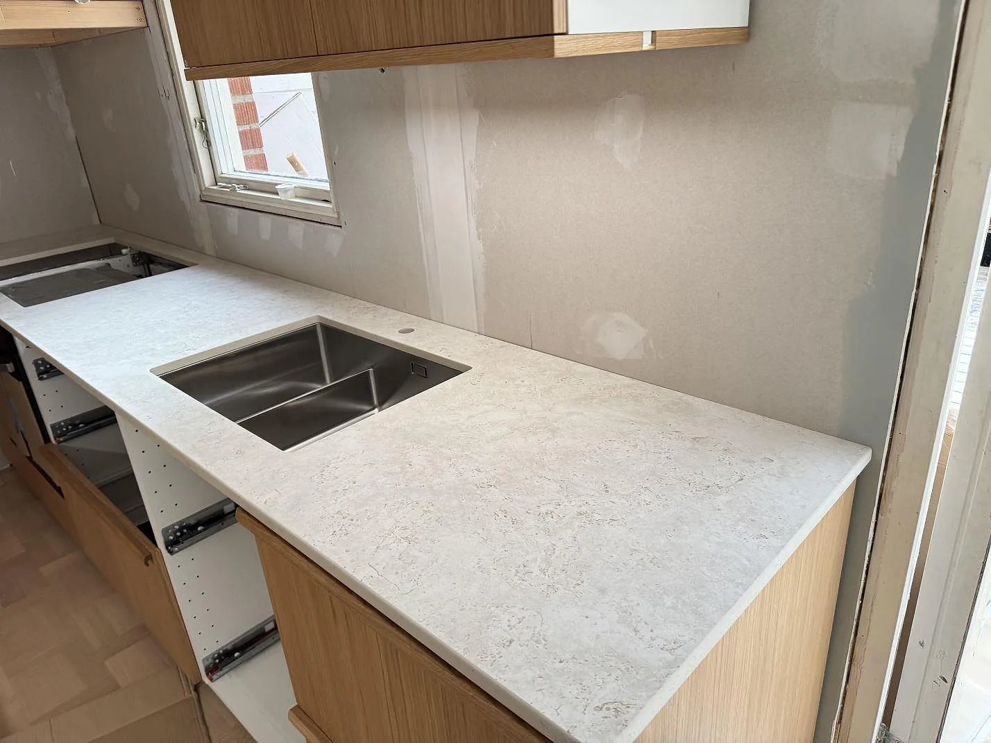 Ljus köksbänkskiva i matt keramik, Dekton Tk05 Sabbia, med rostfri diskho och blandarhål under montering i Tollered.