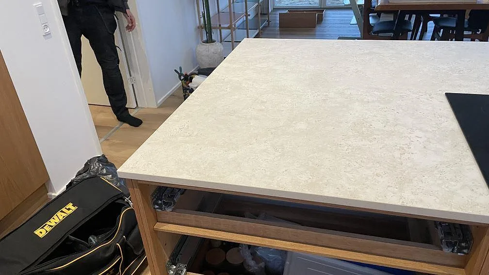 Köksö i matt Dekton Tk05 Sabbia med bred arbetsyta och öppna trästommar under montering i Vejle.