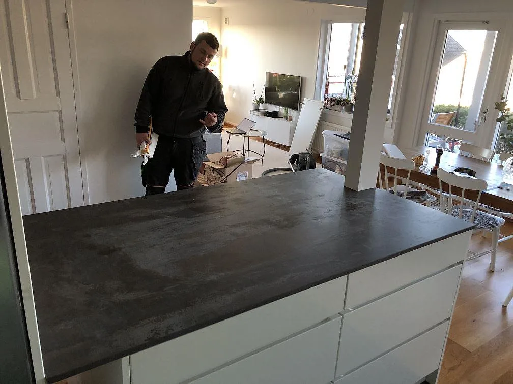 Köksbänkskiva i Dekton Trilium, matt, på en köksö med vita lådfronter i ett öppet kök i Billdal, mörk oxiderad metallgrå ton.