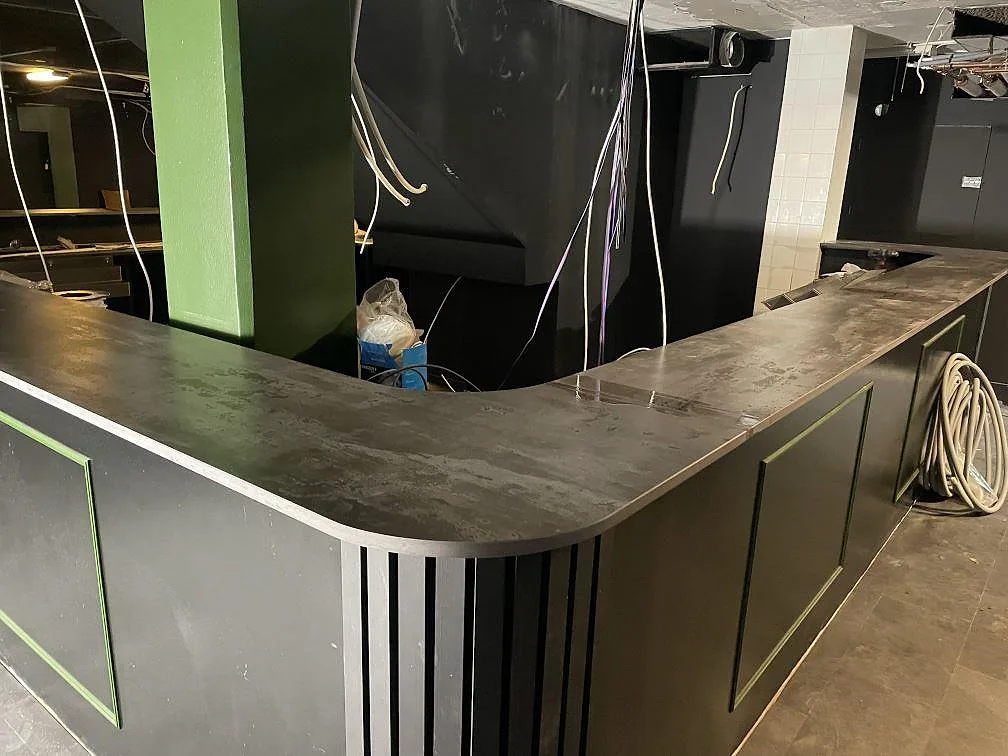 Lång bardisk i matt Dekton Trilium keramik runt gröna pelare och svart slätad bas i en restauranglokal i Borås.
