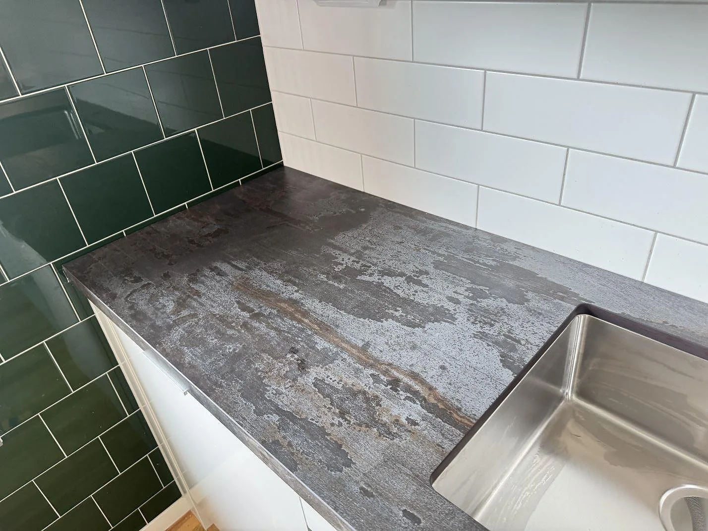Köksbänkskiva i matt Dekton Trilium med rostfri diskho vid grön och vit kaklad hörnvägg i Malmö.