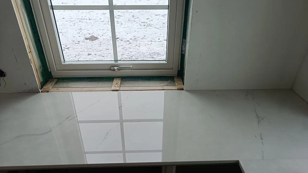 Polerad vit Dekton Tundra bänkskiva under fönstret med grå ådring i ett kök under montering i Borås.