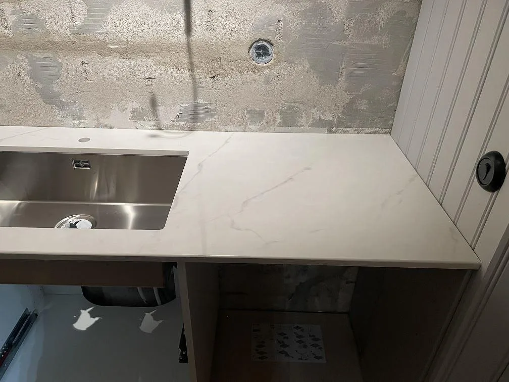 Köksbänkskiva i ljus Dekton Tundra med underlimmad rostfri diskho mot oputsad vägg i ett kök under montering i Rydebäck.