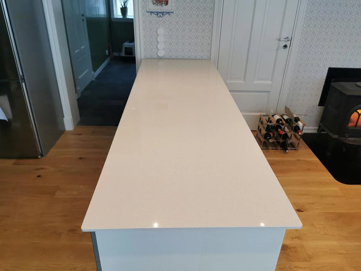 Köksö med bänkskiva i ljus krämvit Dekton Vienna keramik, polerad, i ett kök med trägolv och vedeldad kamin i Kungshamn.