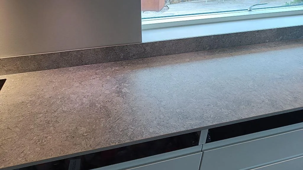Lång arbetsyta i matt Dekton VK03 Grigio med låg stänkkant under fönster i ett kök i Kullavik.