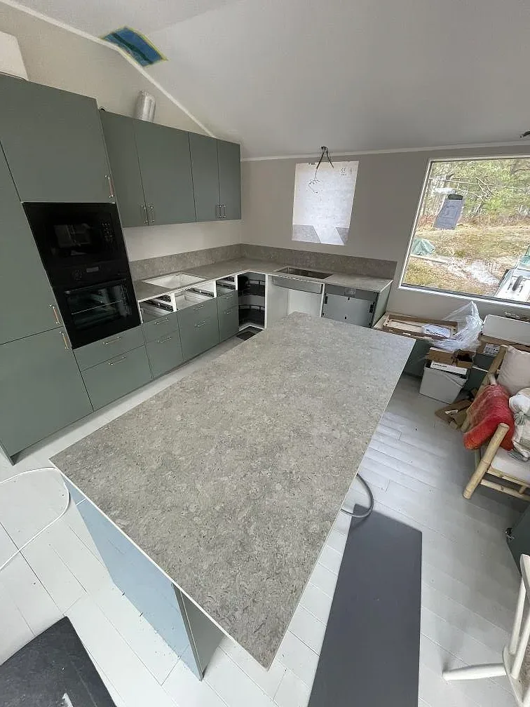 Köksbänkskiva i grå Dekton VK03 Grigio, matt keramik med marmormönster, på en köksö med salviagröna luckor i ett kök under montering i Värmdö.