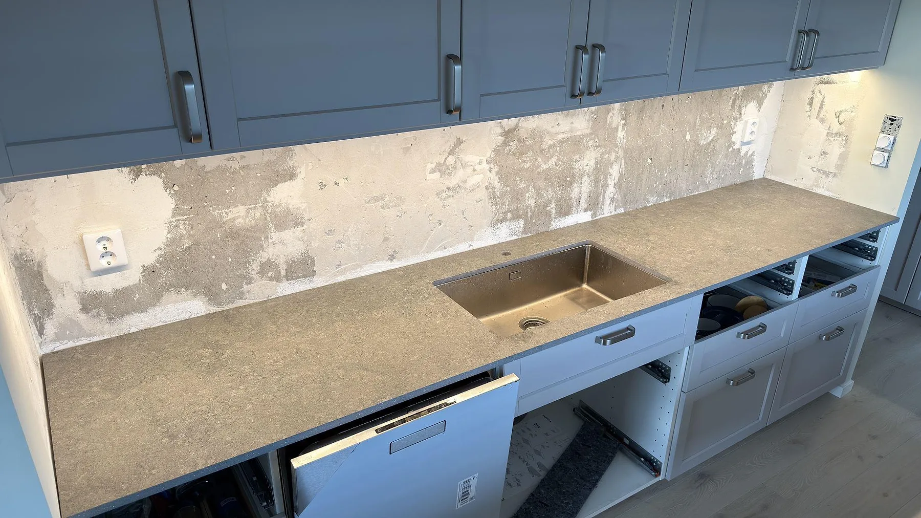 Bänkrad i matt Dekton Vk04 Grafite med rostfri diskho, grå keramik och ofärdig väggyta i ett kök i Lidingö.