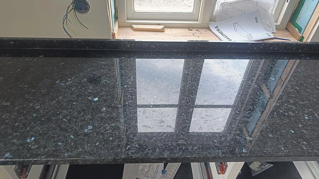 Närbild på polerad svart Emerald Pearl granit med uppvikt stänkkant och fönsterreflex i Hönö.