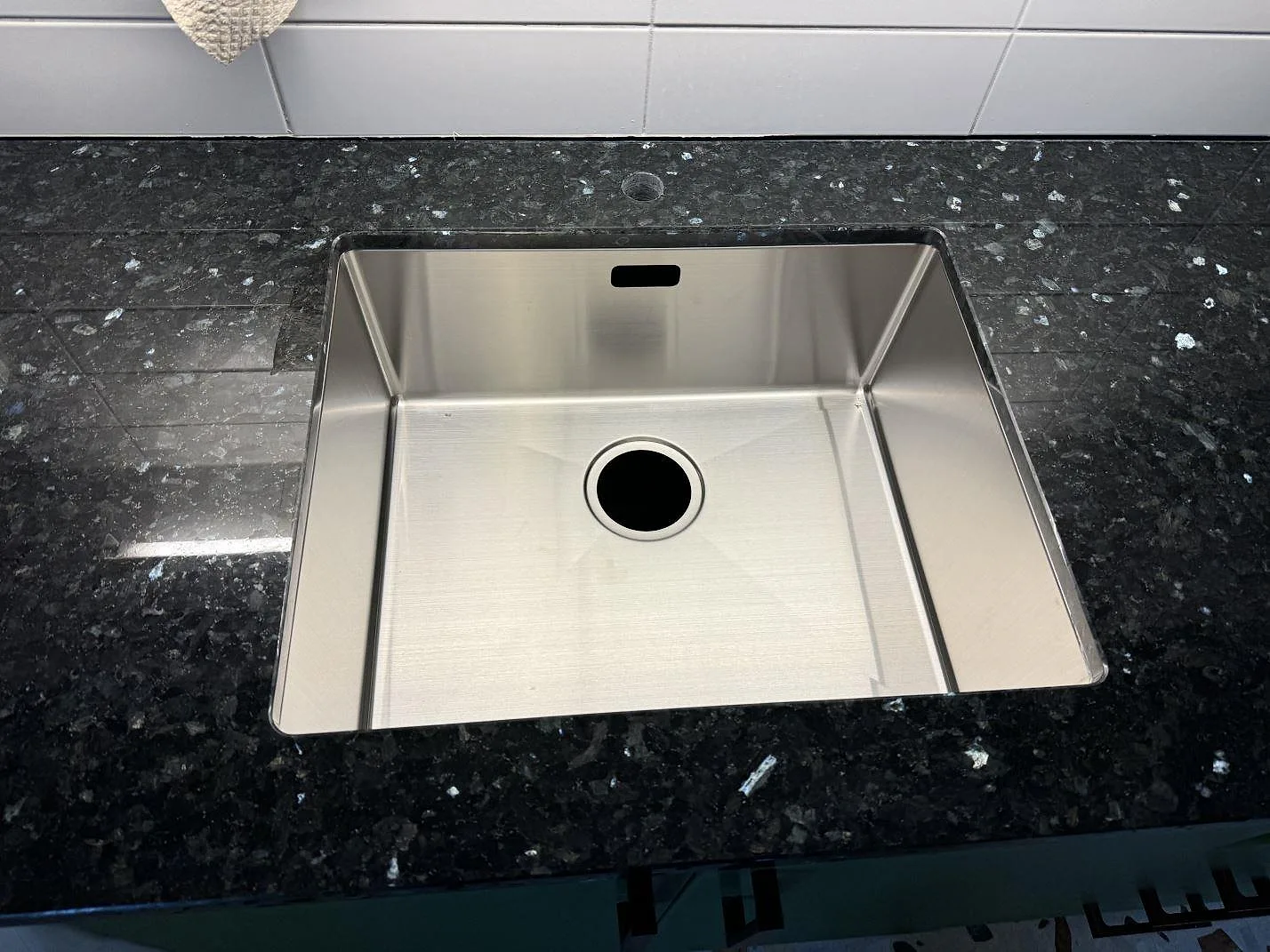 Närbild av underlimmad rostfri diskho i polerad Emerald Pearl granit med vit kakelvägg i ett kök i Lund.
