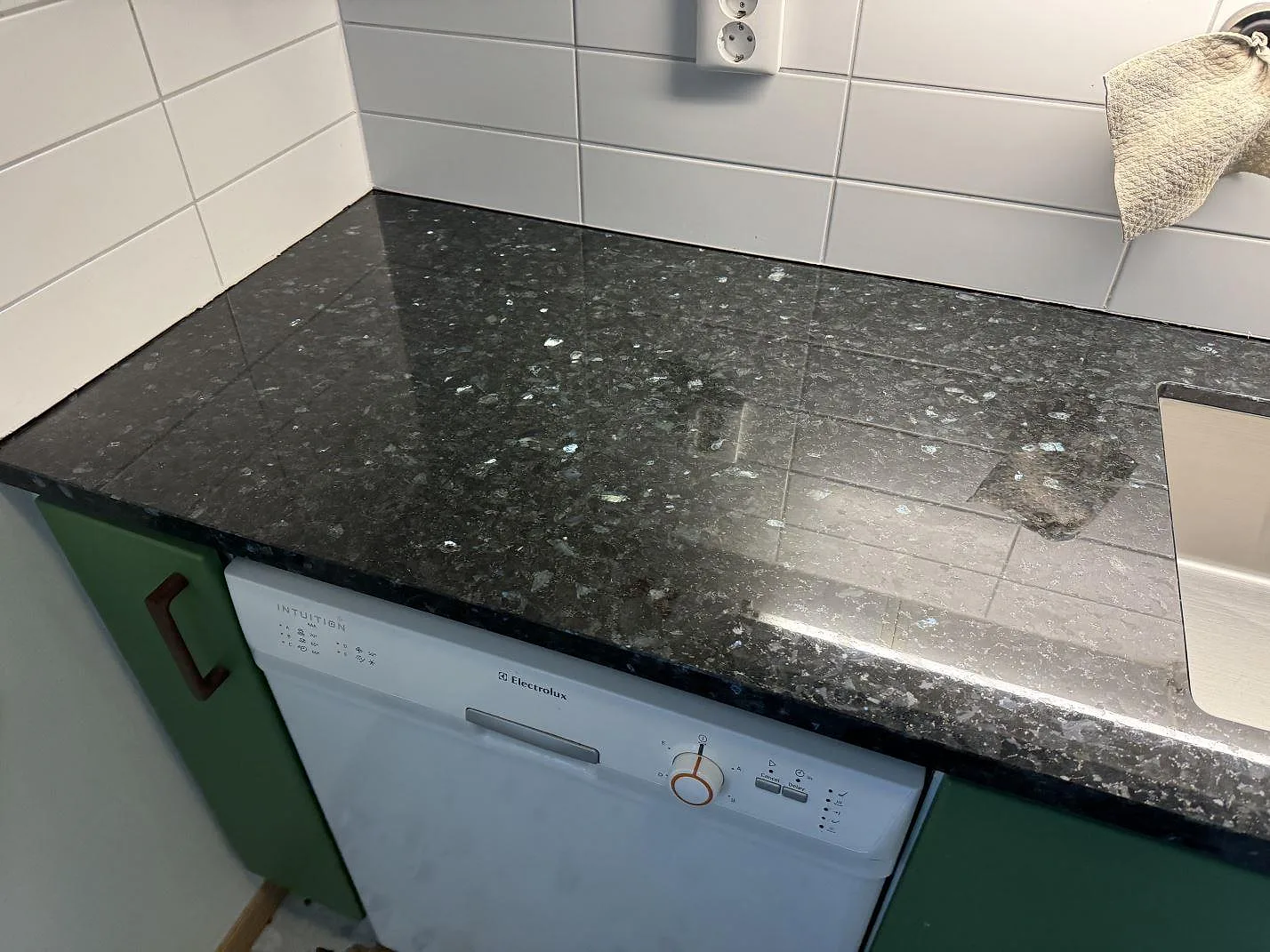 Polerad köksbänkskiva i Emerald Pearl granit över diskmaskin och gröna luckor i ett kök i Lund.