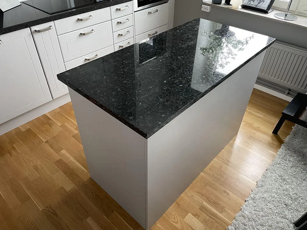 Köksö med polerad bänkskiva i Emerald Pearl granit, mörk natursten med blågröna skiftningar, i ett vitt kök i Tyreso.