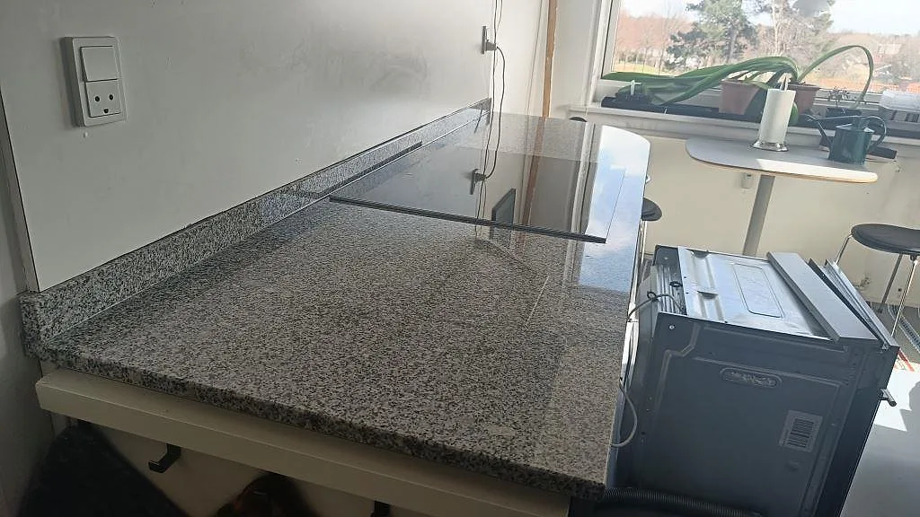 Köksbänkskiva i grå granit Grigio Cristallo, matt yta, installerad längs väggen i ett kök i Gentofte med fönster och utsikt i bakgrunden.