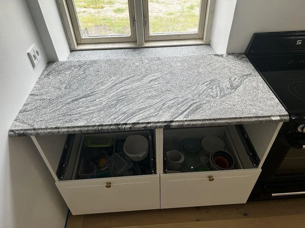 Installerad köksbänkskiva i polerad Grigio Cristallo granit under fönster, med vita underskåp och spis intill i Kirke Hyllinge.