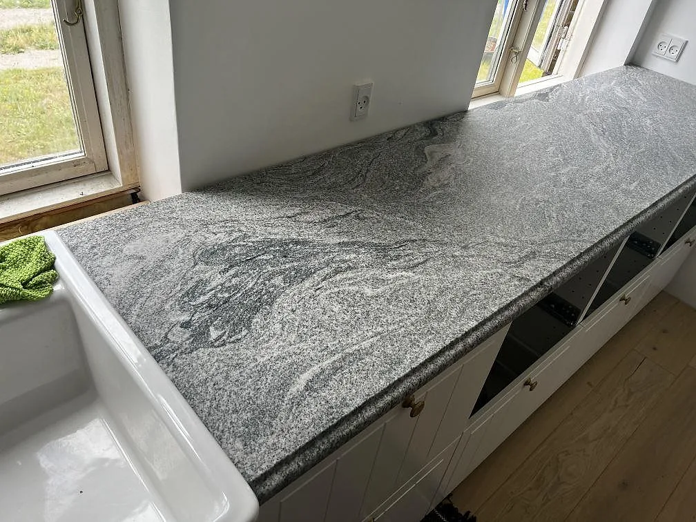 Köksbänkskiva i polerad Grigio Cristallo granit vid vit diskho, fönster och vita skåpstommar i Kirke Hyllinge.