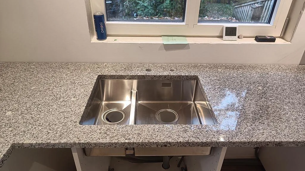 Underlimmad rostfri dubbelho i en polerad Grigio Cristallo granitskiva vid fönstret i ett kök i Laholm.