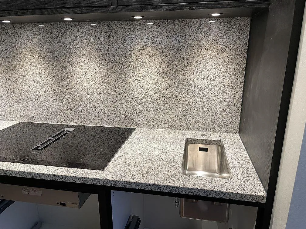 Väggsida med polerad Grigio Cristallo granit, stänkskydd, häll och rostfri diskho under LED-belysning i Simrishamn.