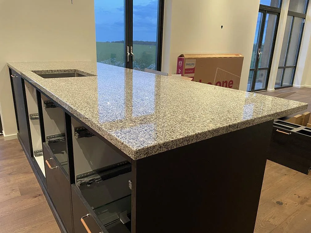 Köksö med polerad bänkskiva i Grigio Cristallo granit, svart köksinredning och stora fönster i ett kök i Simrishamn.