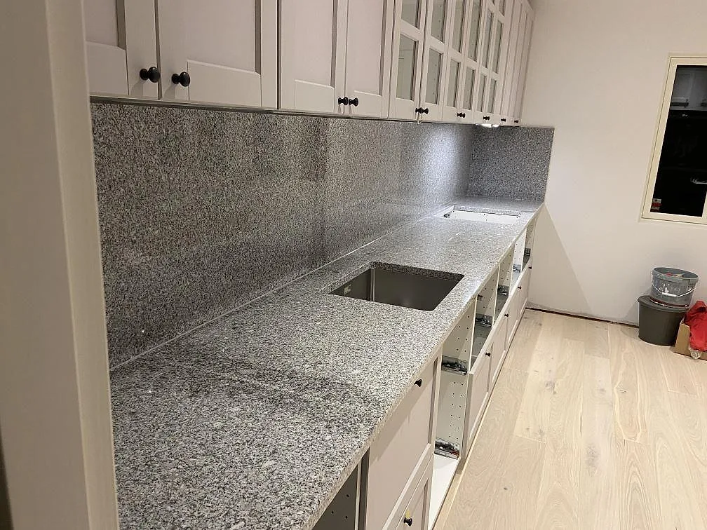 Köksbänkskiva i polerad Grigio Cristallo granit med uppvikt stänkskydd i samma sten, underlimmad diskho och ljusa köksluckor i ett kök i Ystad.