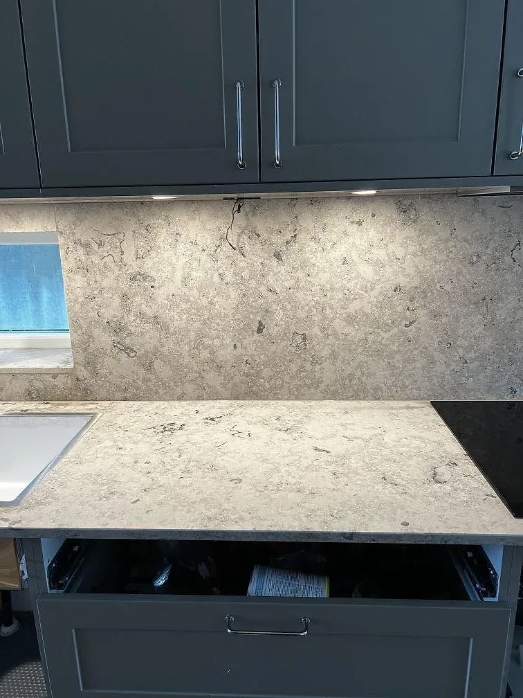 Hög väggbeklädnad och bänkskiva i matt Jura Grey kalksten mellan mörkgrå köksstommar i Bankeryd.