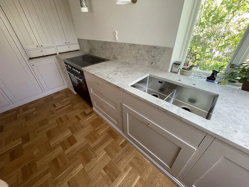 Närbild på köksbänkskiva i matt Jura Grey med rostfri diskho och stänkkant under fönster i Bromma.