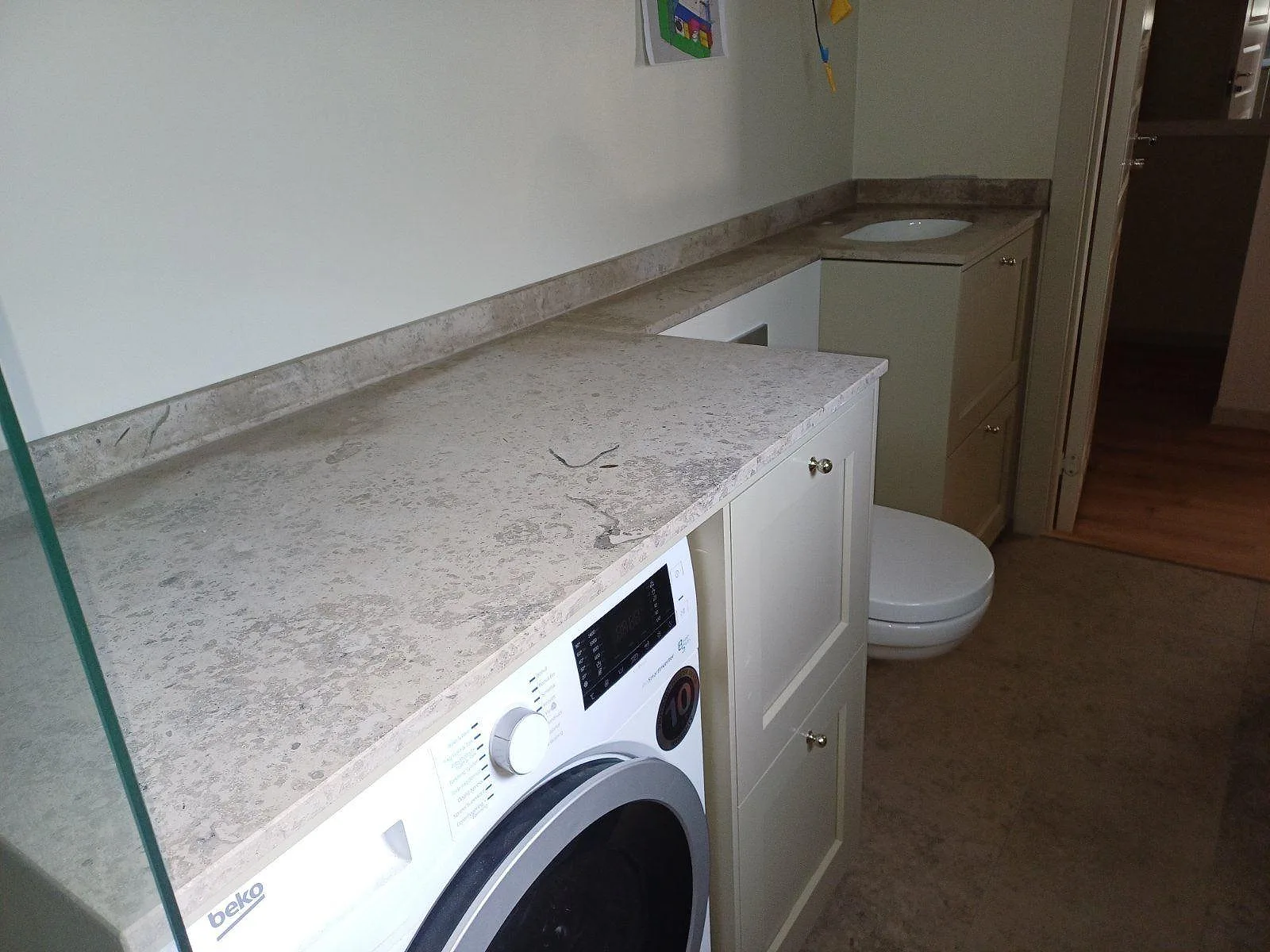 Bänkskiva i matt Jura Grey, 20 mm, som grå kalkstensyta över tvättmaskin och vit badrumsinredning i Ingarö.