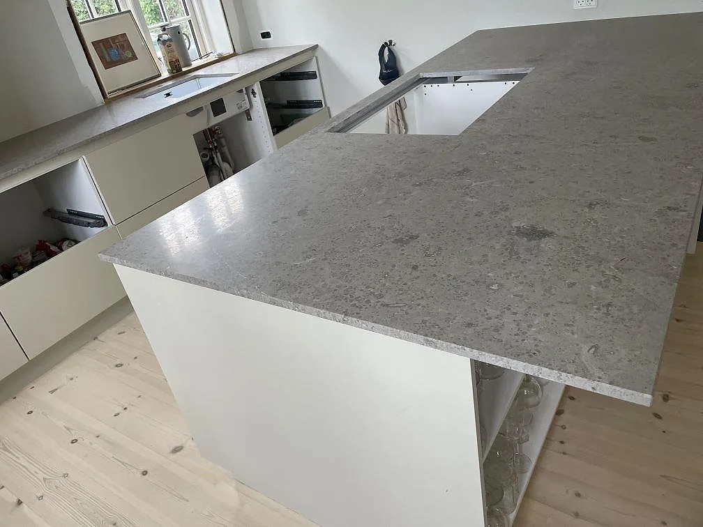 Närbild av köksö i matt Jura Grey kalksten med gråbeige fossilstruktur, vit köksinredning och fönster i Kongens Lyngby.