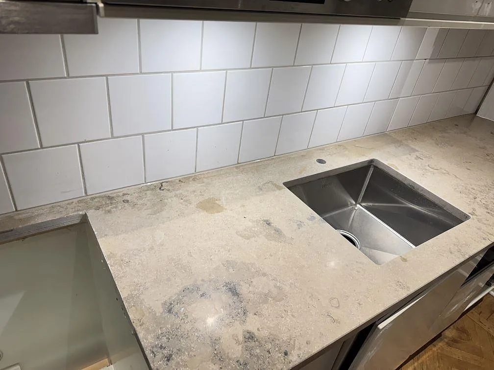 Matt köksbänkskiva i Jura Grey kalksten i Lund, sedd uppifrån med lång arbetsyta, diskho och öppet urtag framför vitt kakel.