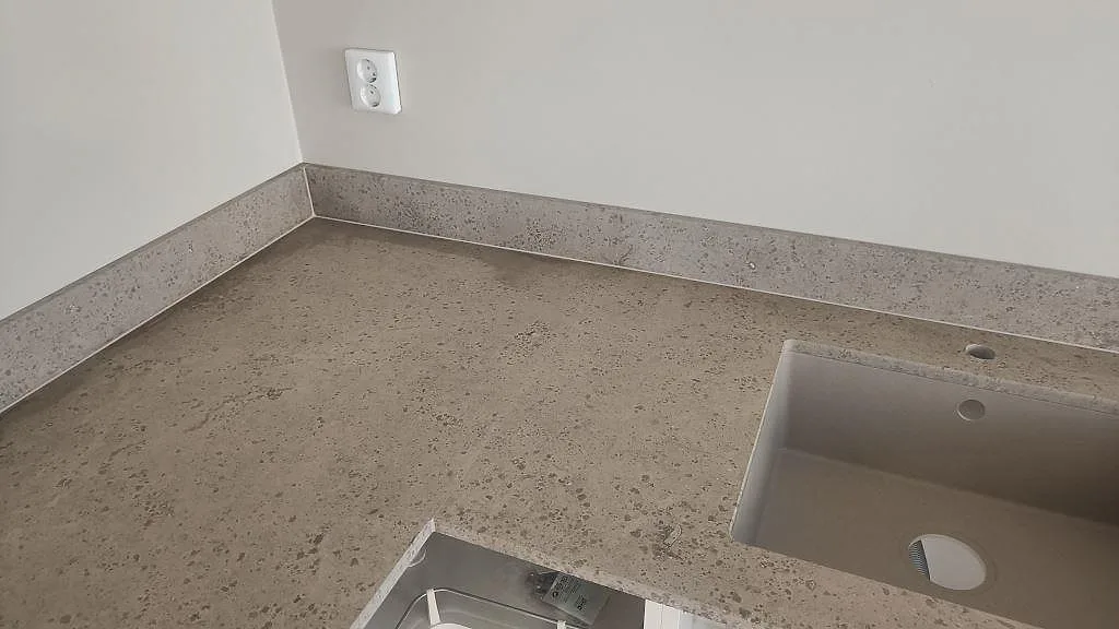 Hörnvy över L-formad köksbänkskiva i matt Jura Grey kalksten med diskho, stänkkant och vit vägg i Malmö.