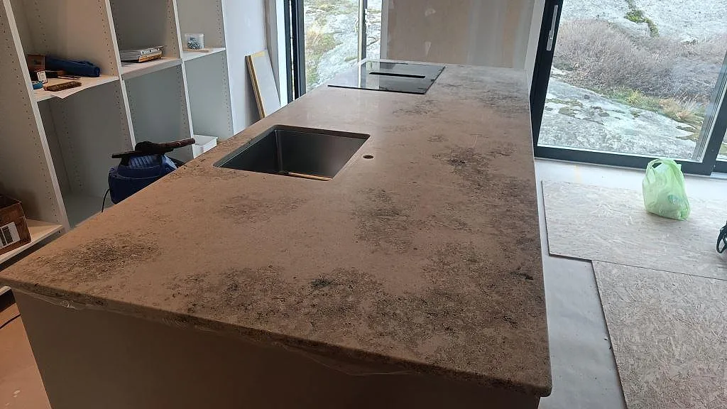 Lång köksö med matt Jura Grey bänkskiva, rostfri diskho och häll vid stora fönster i Marstrand.