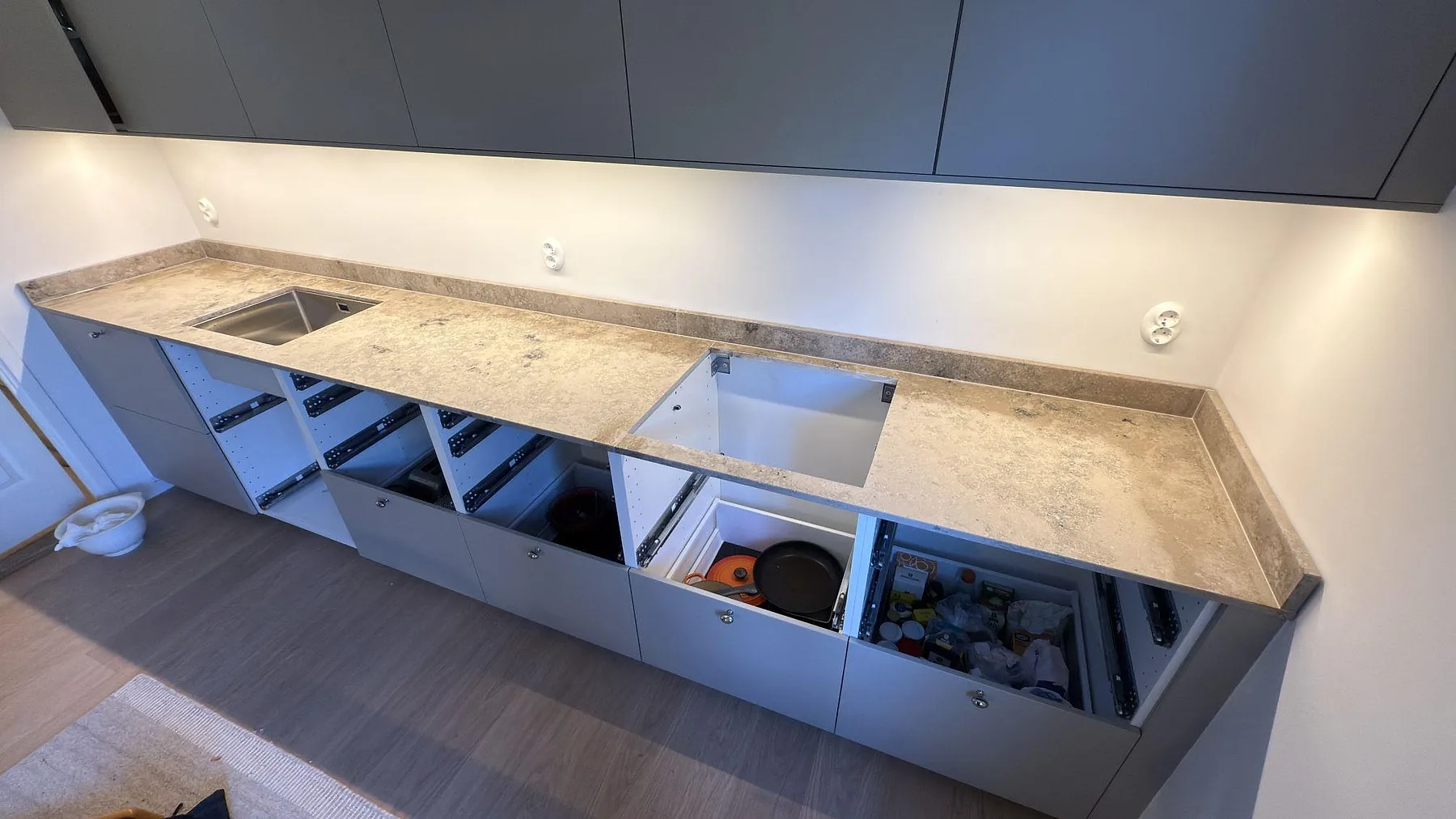 Lång köksbänkskiva i matt Jura Grey med rostfri diskho, hällöppning och öppna köksstommar under montering i Saltsjö Boo.