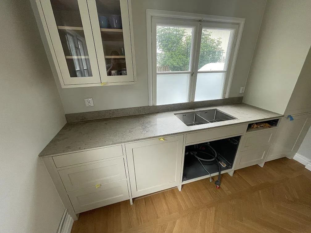 Köksbänkskiva i matt Jura Grey kalksten vid fönster, med diskho och låg stänkkant i ett kök i Stockholm.