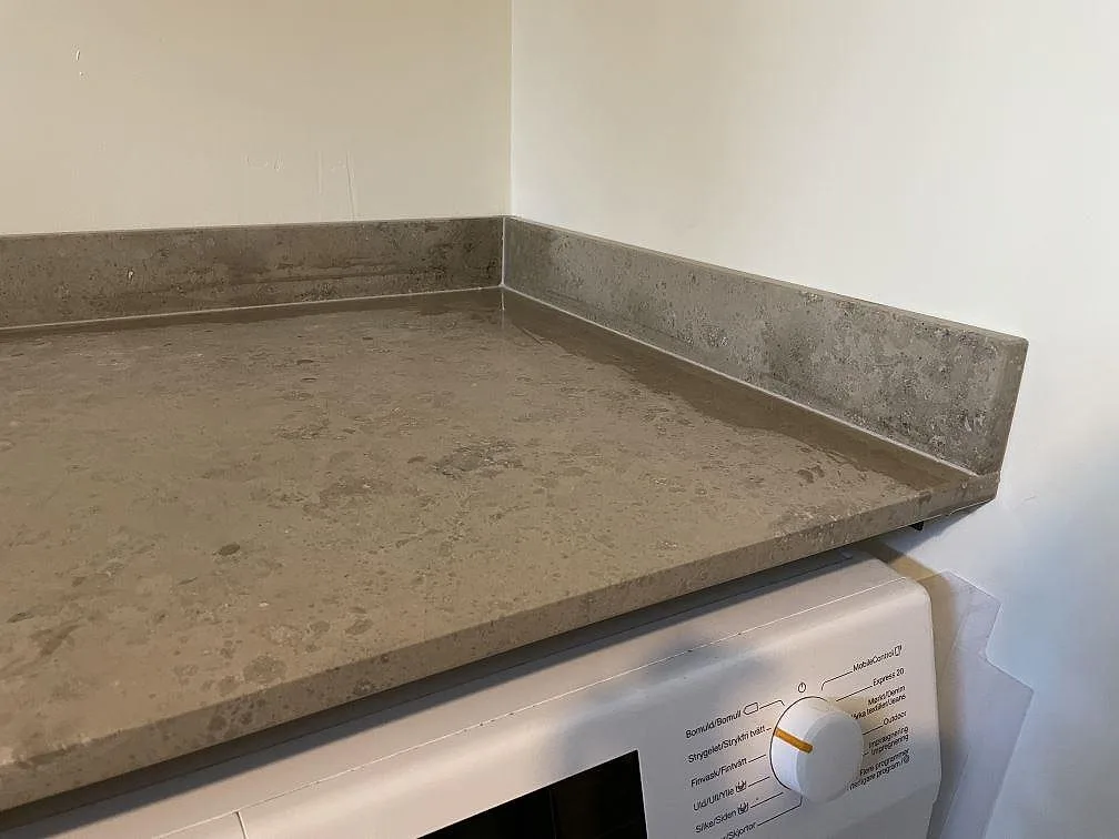 Detalj av matt Jura Grey kalksten med uppvikt stänkkant ovanför Miele tvättmaskin i en tvättstuga i Töllsjö.