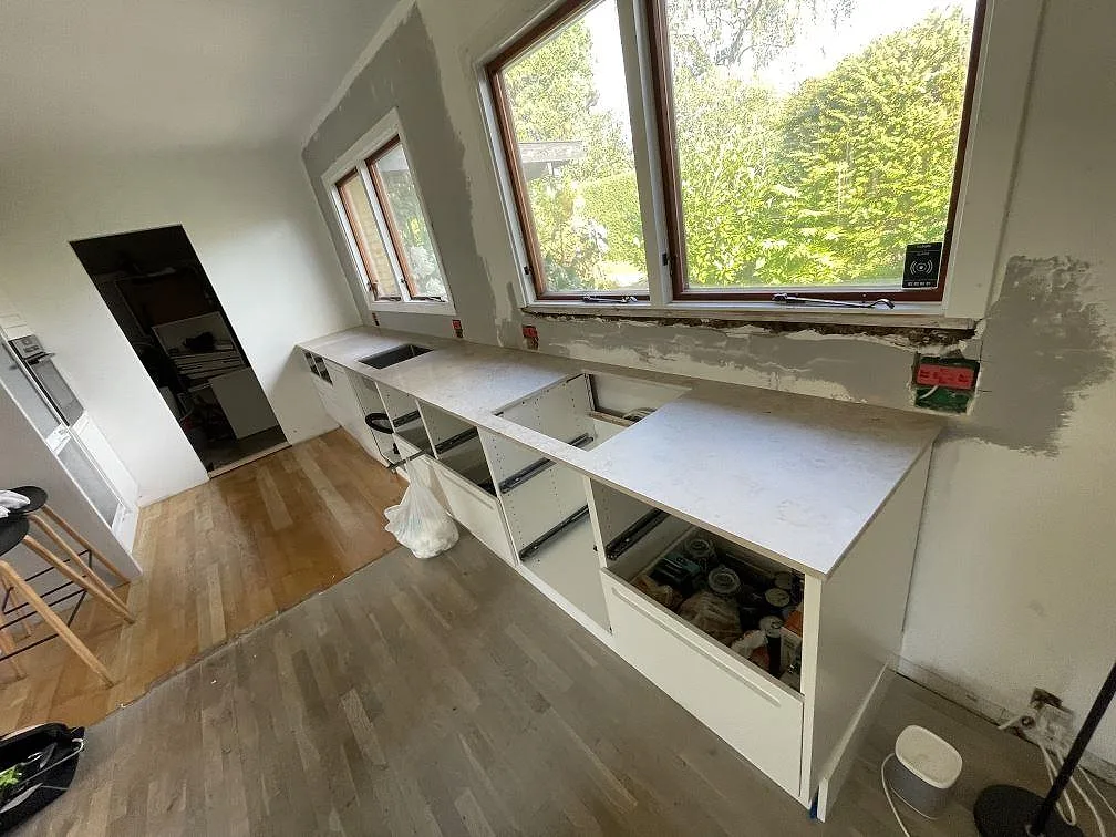 U-format kök i Vedbæk med köksbänkskivor i matt Jura Grey kalksten, vita stommar, stora fönster och trägolv under montering.