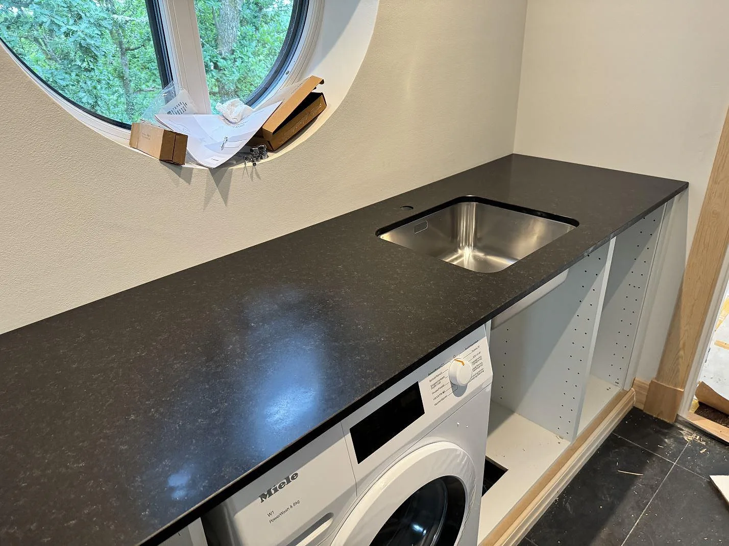 Bänkskiva i matt Nero Assoluto granit med underlimmad rostfri diskho, rund fönsteröppning och tvättmaskin i Alingsås.