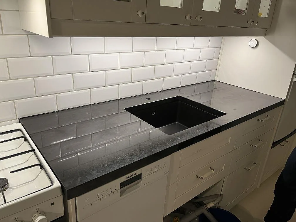 Köksbänkskiva i svart Nero Assoluto granit, polerad, med svart ovanpåmonterad diskho och vitt kakel i ett kök i Bromma.