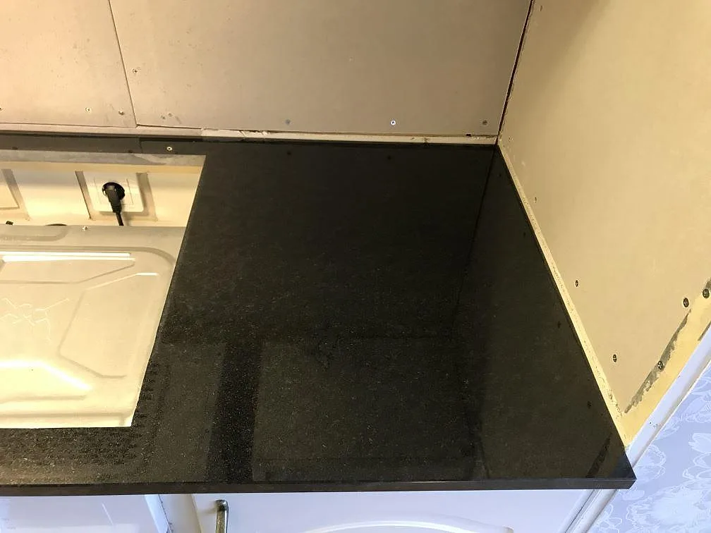 L formad köksbänkskiva i polerad Nero Assoluto granit med urtag för häll, installerad i situ mot omålade väggar i ett kök i Falköping.