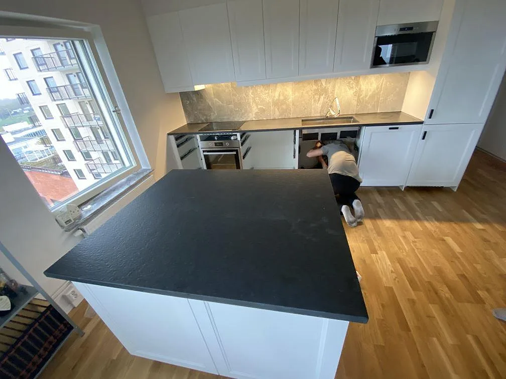 Köksbänkskiva och köksö i matt Nero Assoluto Flamed and Brushed granit under montering i ett vitt kök i Limhamn.