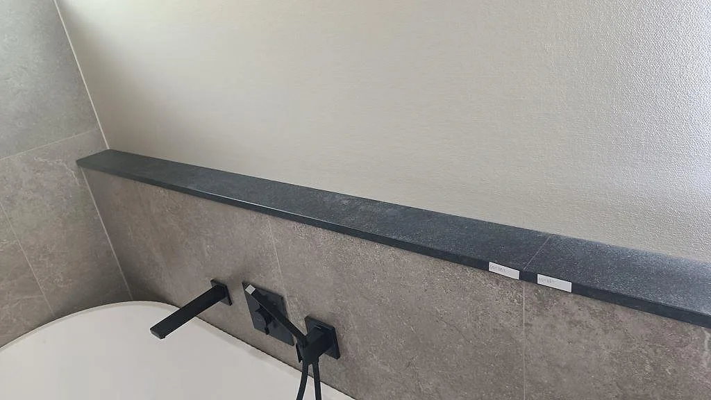 Matt hyllplan i Nero Assoluto Flamed and Brushed granit ovanför badkar med grå väggplattor i Mölndal.