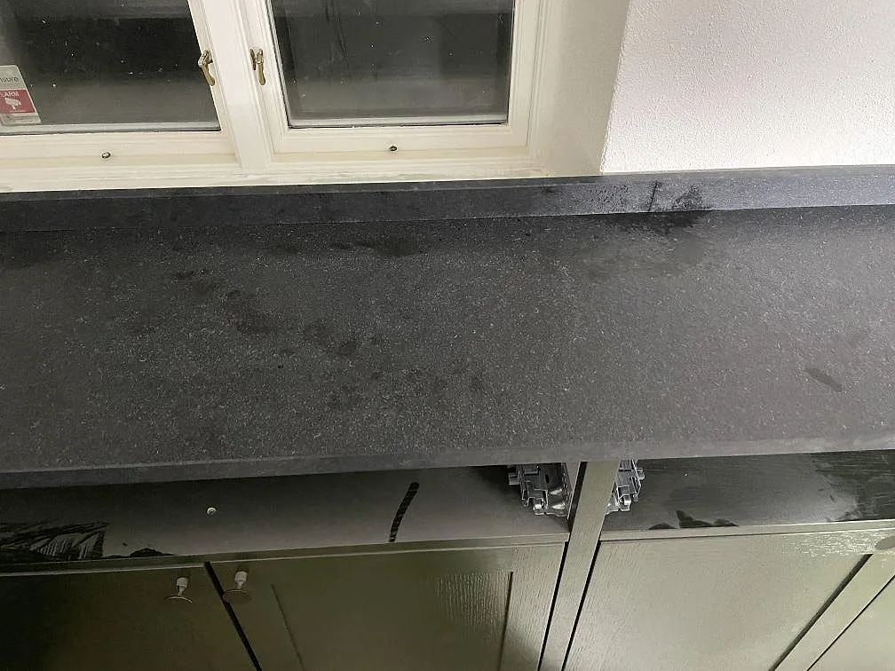 Köksbänkskiva i svart granit Nero Assoluto Flamed and Brushed med matt lädrad finish vid fönstret i ett kök i Rydsgaard.