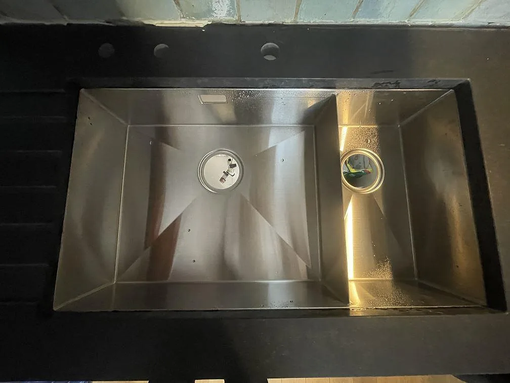 Underlimmad rostfri diskho med stort och litet tråg i en matt svart Nero Assoluto bänkskiva, med tre borrade hål för blandare, i ett kök i Frövi.