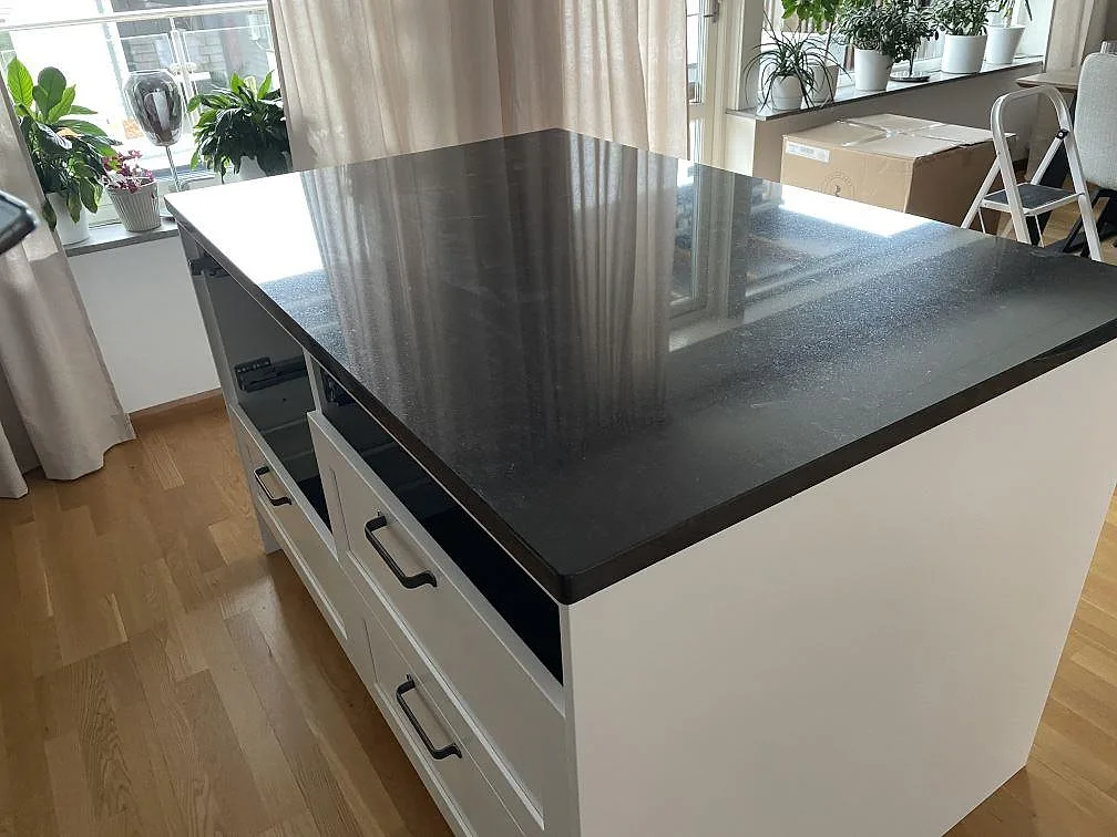 Köksöskiva i svart Nero Assoluto granit, polerad, på vit köksö med trägolv och fönsterljus i Huskvarna.
