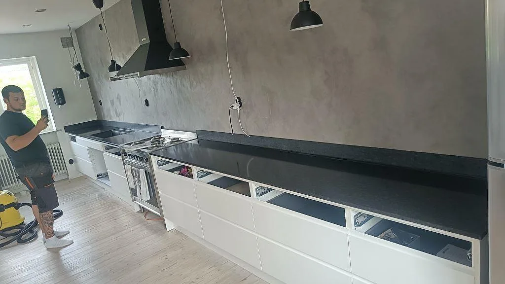 Köksbänkskiva i svart Nero Assoluto, matt granit, längs en lång enradsvägg med vita greppfria underskåp och svart fläktkåpa i ett kök i Köpingebro.