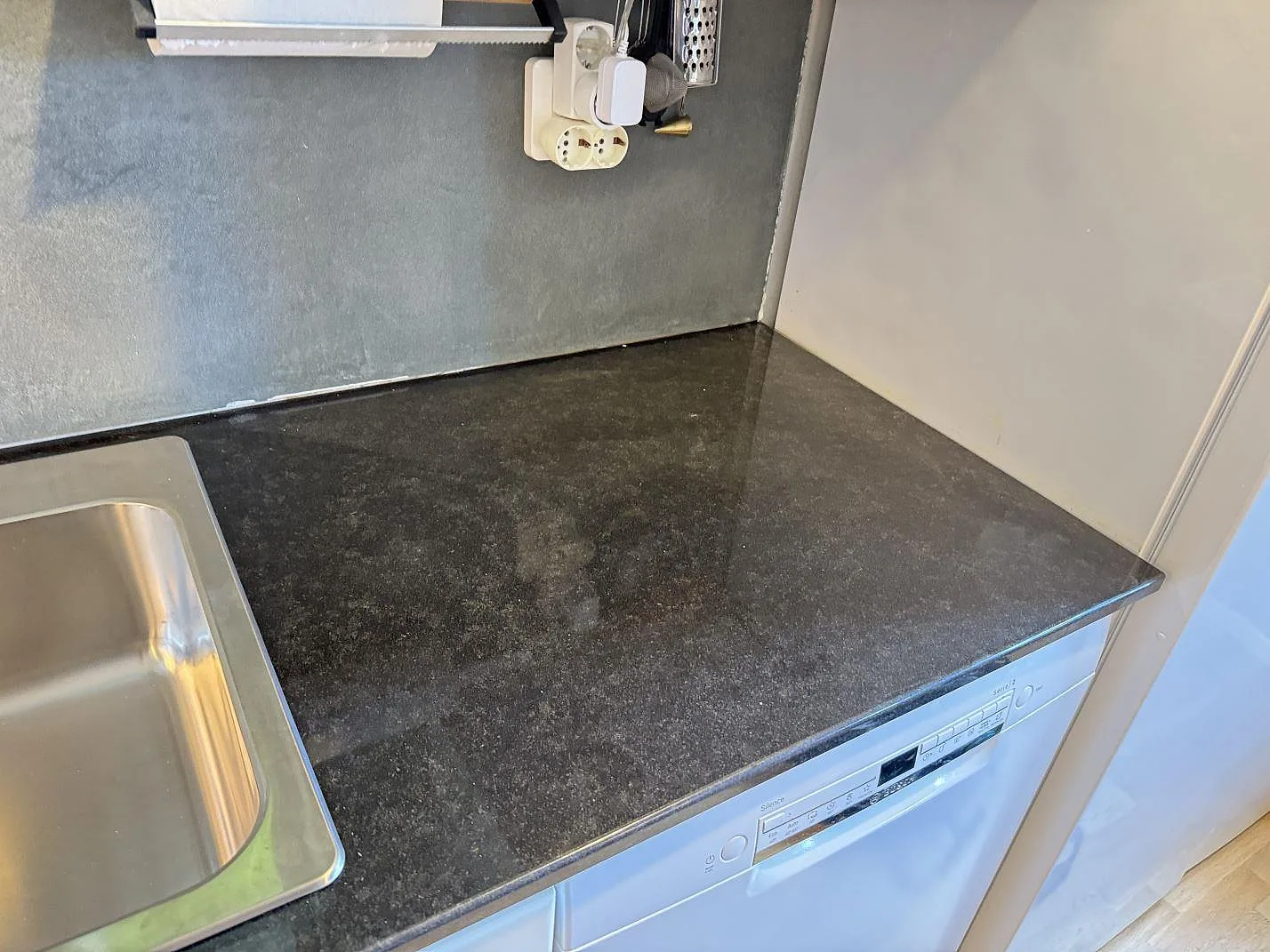 Köksbänkskiva i polerad Nero Assoluto granit med rostfri diskho, diskmaskin och mörk stenyta i ett kök i Kristianstad.