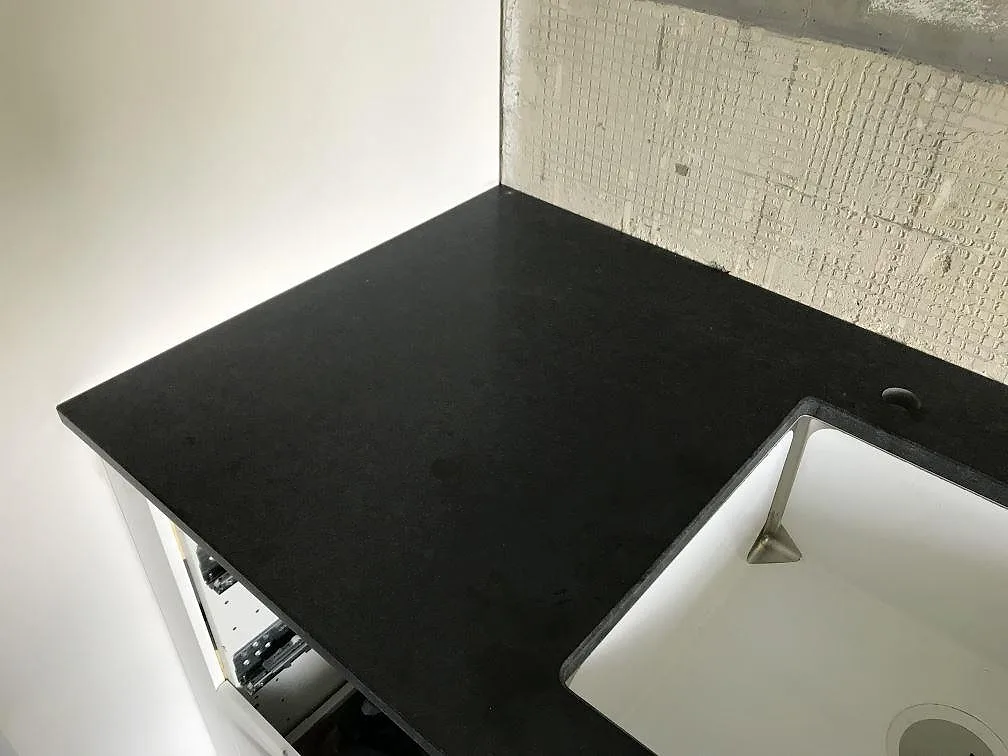 Matt svart Nero Assoluto-granit som vänder in i hörnet i ett kök i Landskrona, med underlimmad vit diskho och öppen stomme under bänkskivan.