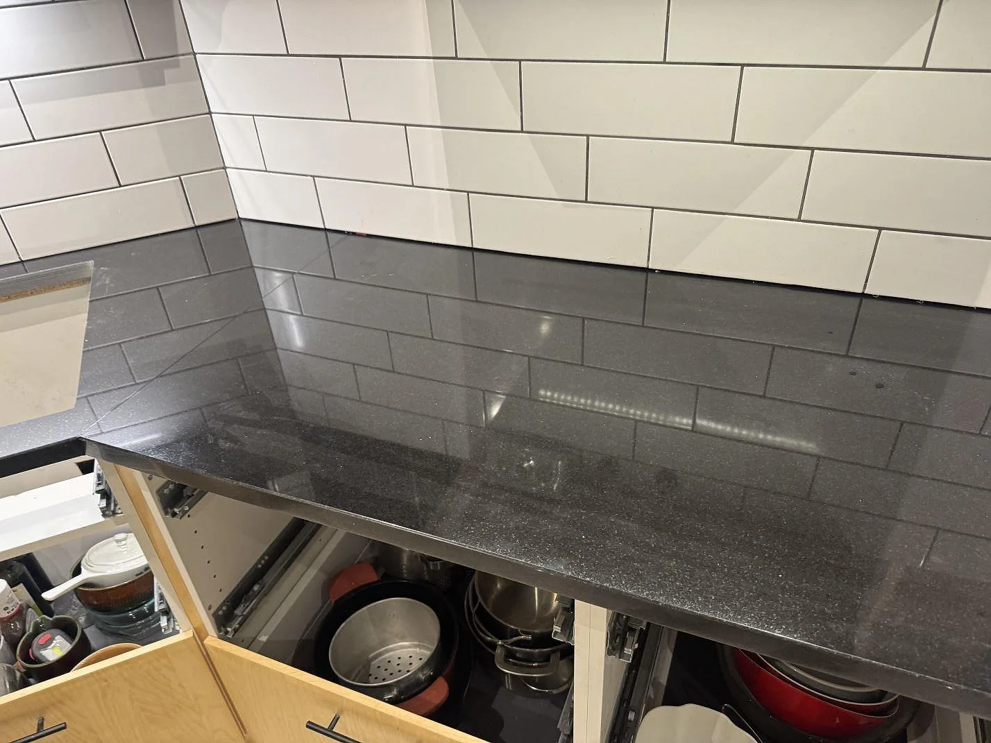 Lång köksbänkskiva i polerad svart Nero Assoluto-granit framför vitt kakel och öppna lådor i Målilla.