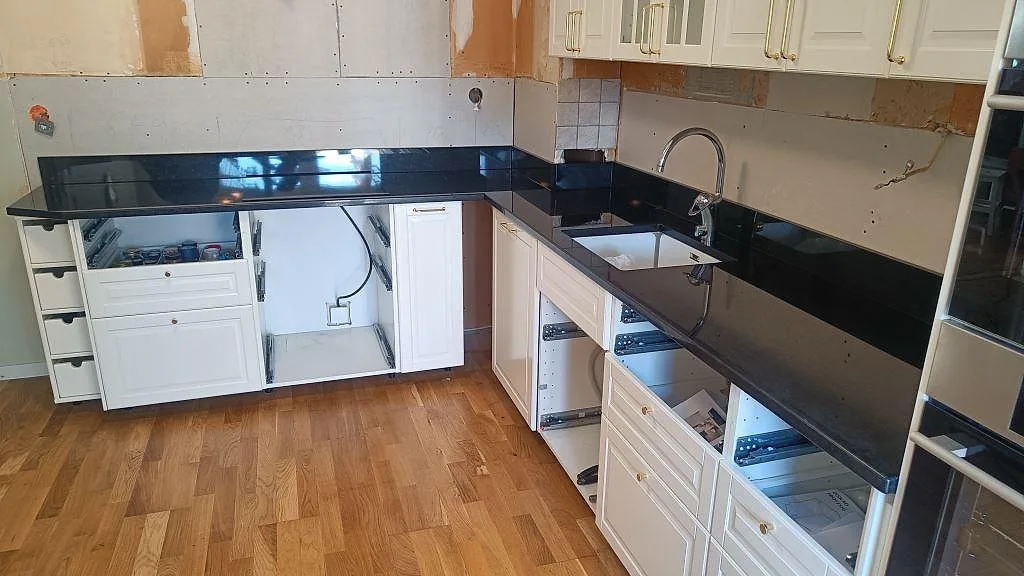 Köksbänkskiva i svart Nero Assoluto-granit, polerad, i L-form med underlimmad diskho och vita köksluckor i ett kök i Malmö.