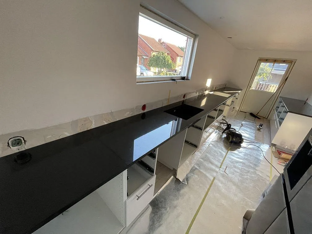 L-formad köksbänkskiva i polerad Nero Assoluto granit med svart diskho, vita skåpstommar och stora fönster i Malmö.