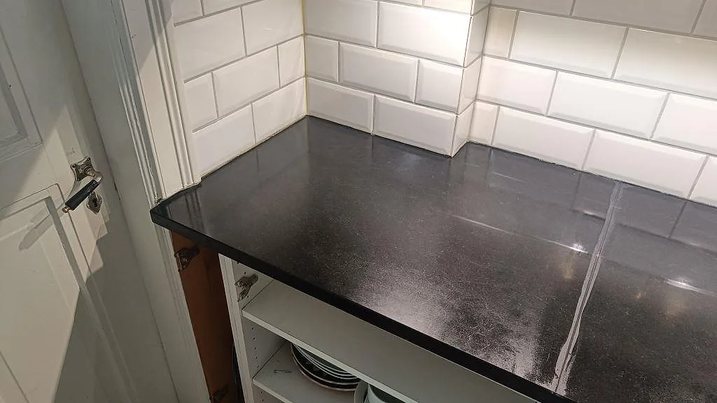 Mörk bänkskiva i matt Nero Assoluto granit mot vit kakelvägg vid hörnet av ett kök i Malmö.