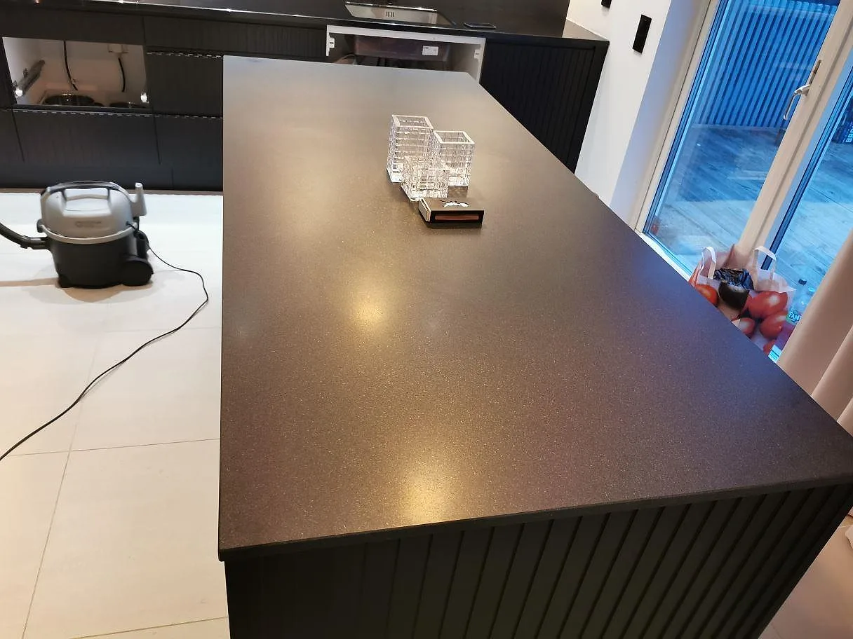 Matt köksbänkskiva i svart Nero Assoluto-granit på en köksö med ribbad svart sockel och stora fönster i ett kök i Skanör.