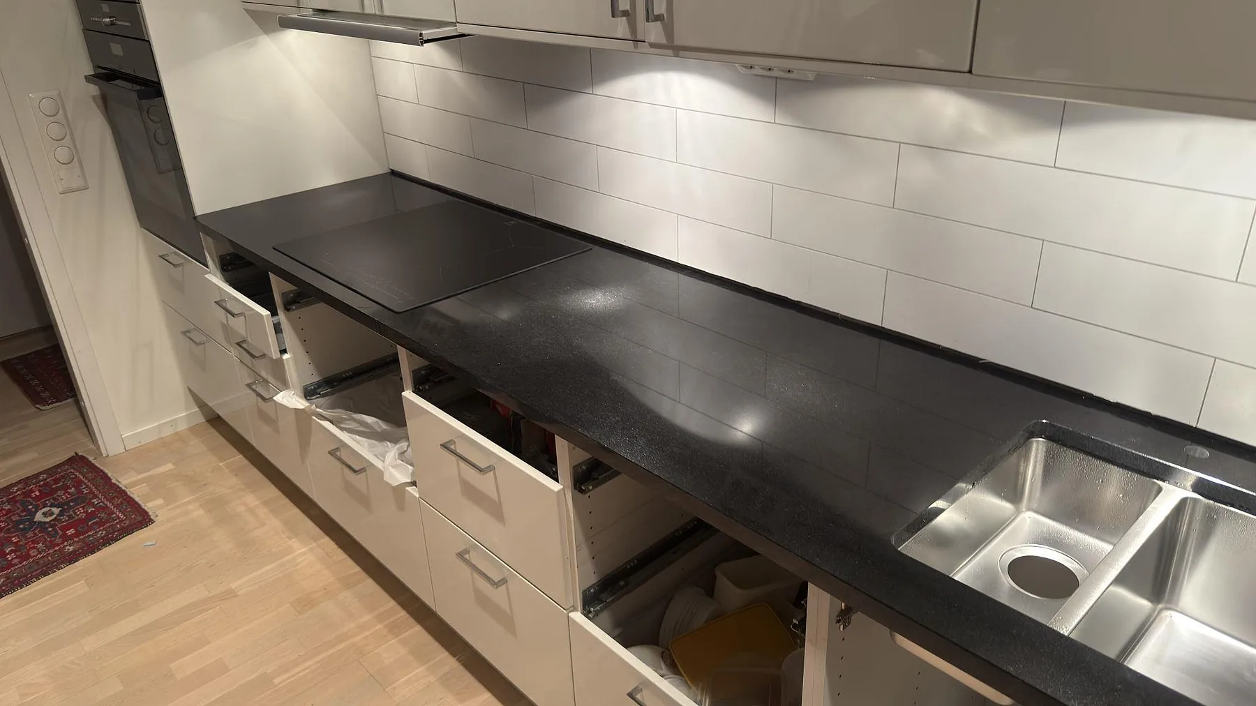 Svart Nero Assoluto granitbänkskiva med infälld induktionshäll och rostfri diskho mot vitt kakel i ett kök i Stockholm.